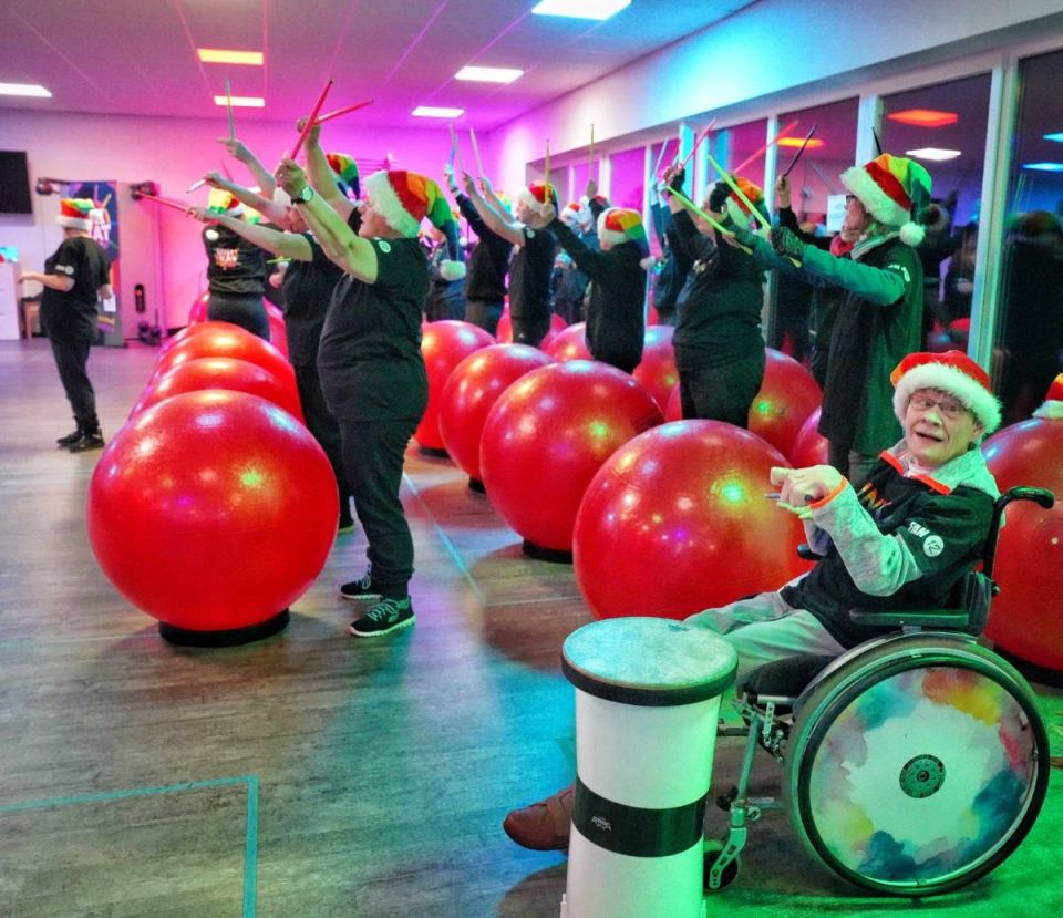 Gruppe trommelt mit Weihnachtsmützen auf roten Bällen, vorne eine Teilnehmerin im Rollstuhl